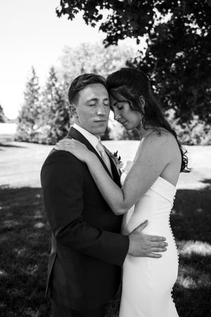 Black and white photo of a bride and groom embracing outdoors, eyes closed, in wedding attire among trees.