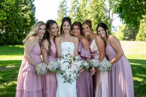 Bride in white gown poses with five bridesmaids in pink dresses holding bouquets outdoors on a sunny day.