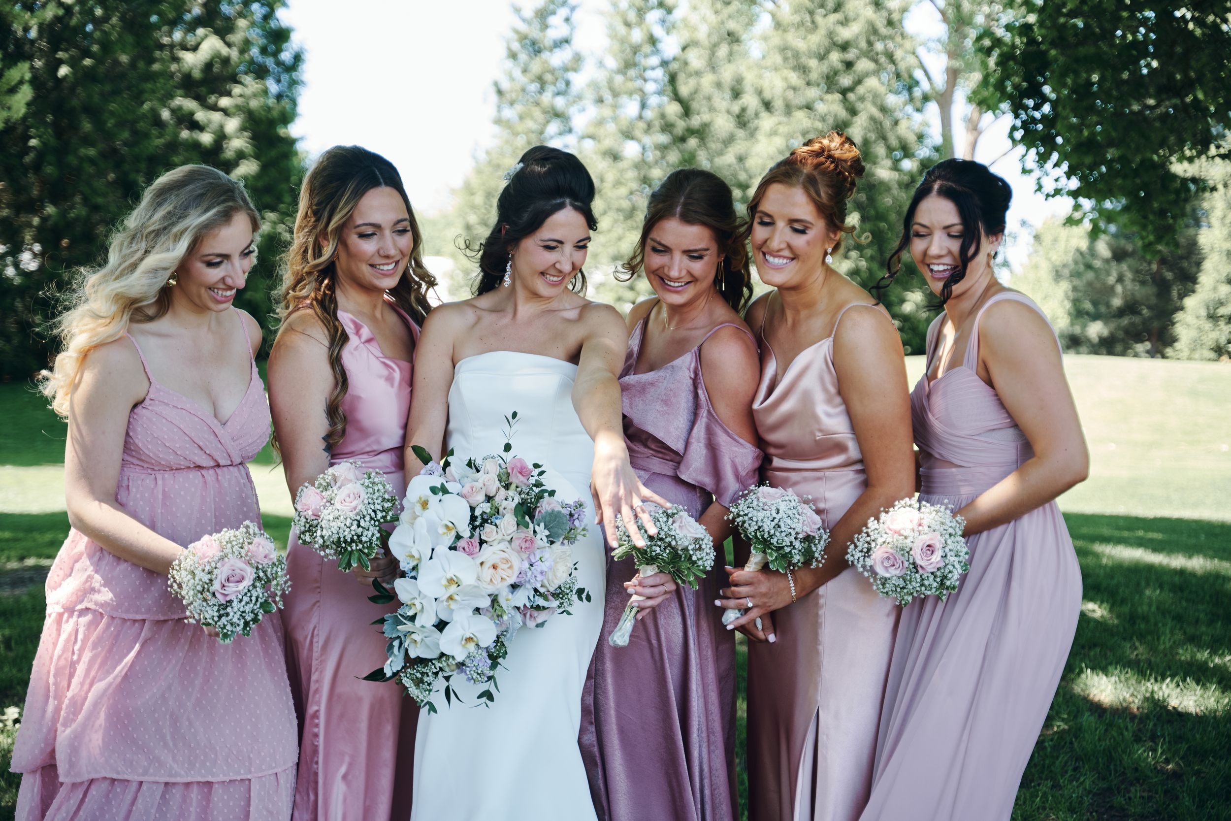 Bride in white gown surrounded by five bridesmaids in pink satin dresses holding floral bouquets outdoors.