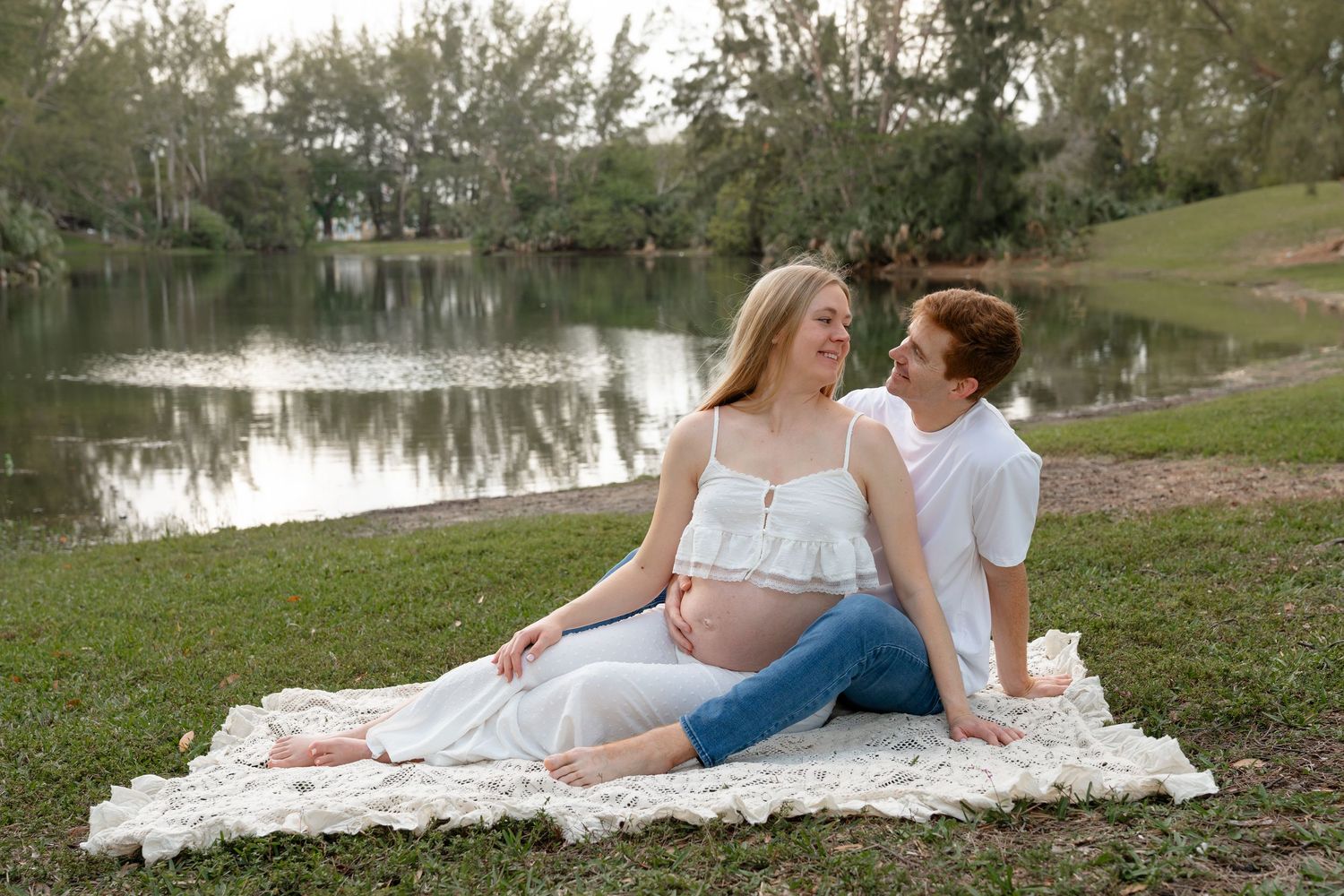Pregnant couple sitting on blanket by serene lake, woman in white dress cradling baby bump, maternity photo session outdoors.