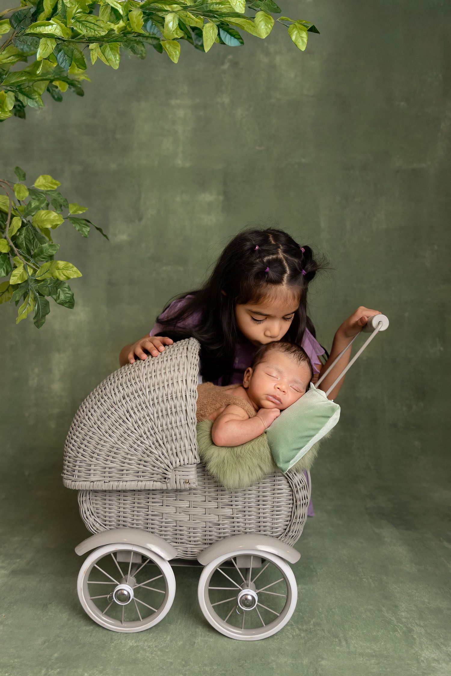 Young girl kissing newborn sibling in vintage wicker pram, green botanical studio backdrop.