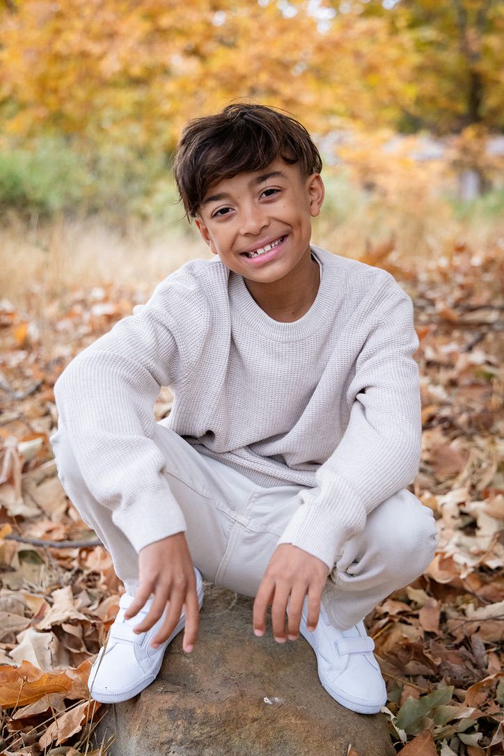 Smiling young boy in beige sweater and pants crouching on a rock surrounded by autumn leaves.
