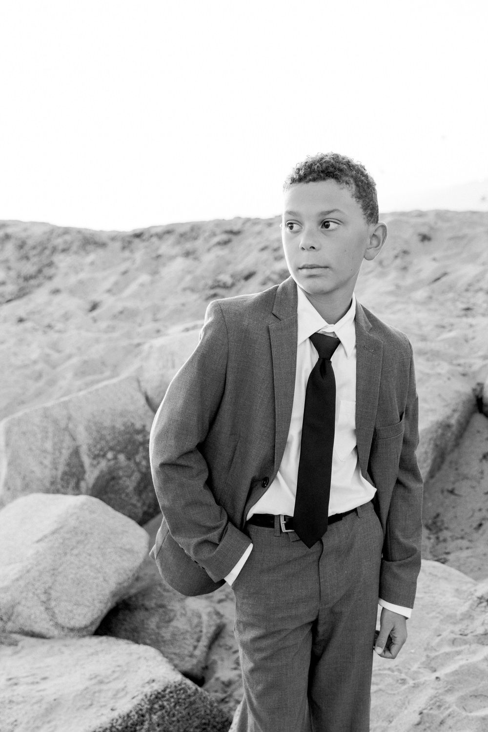 Black and white portrait of a young boy in a suit and tie standing among rocks outdoors.