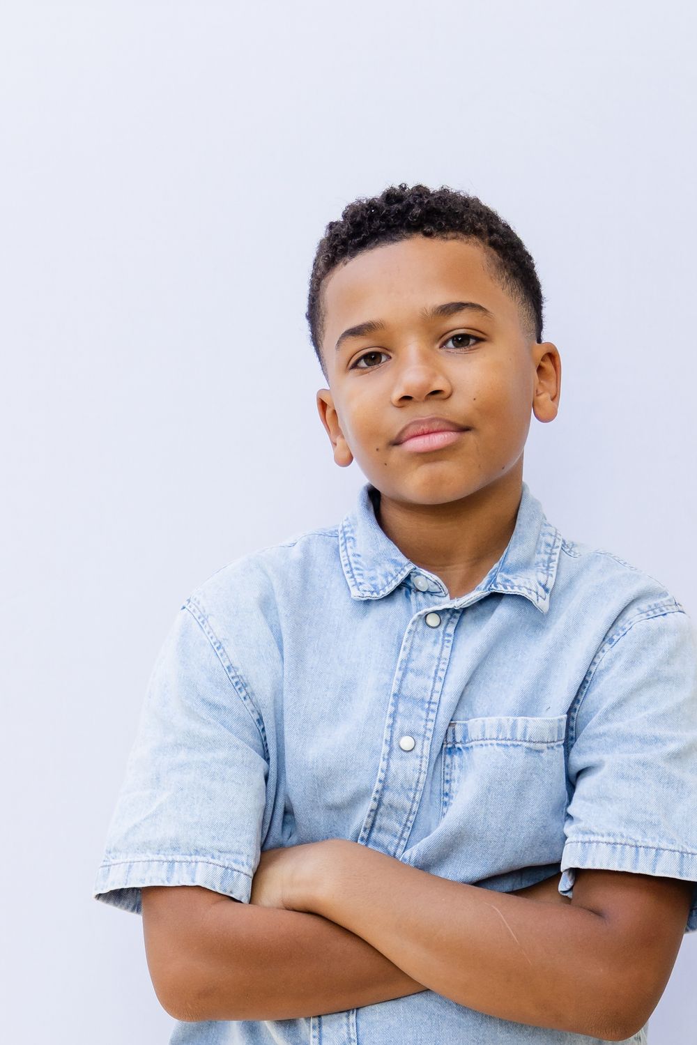 Confident young Black boy in a denim shirt stands with arms crossed against a white background.