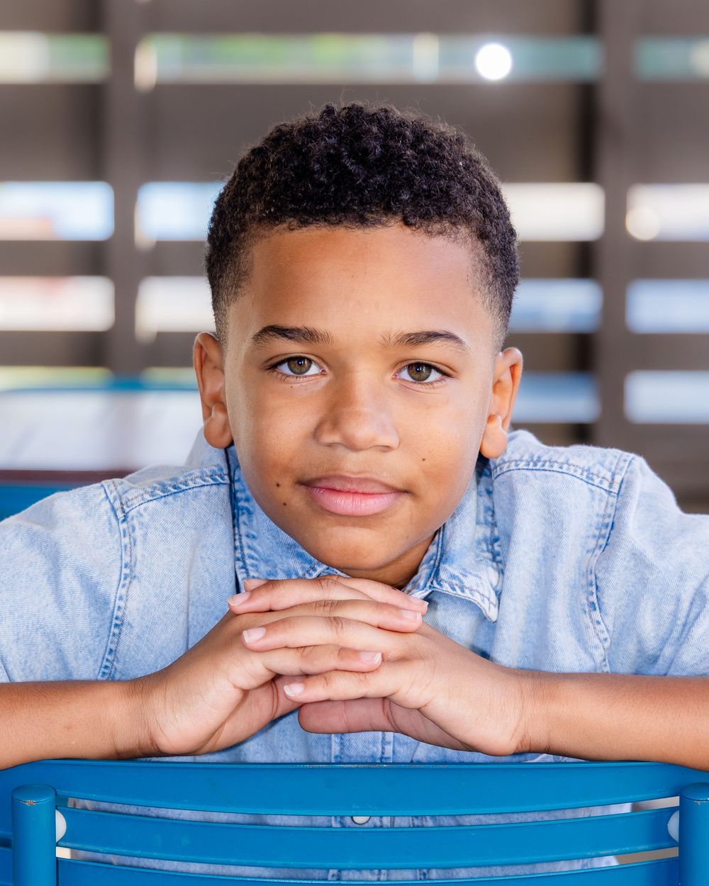 A young boy in a denim shirt rests his chin on clasped hands, smiling at the camera while seated at a blue chair.