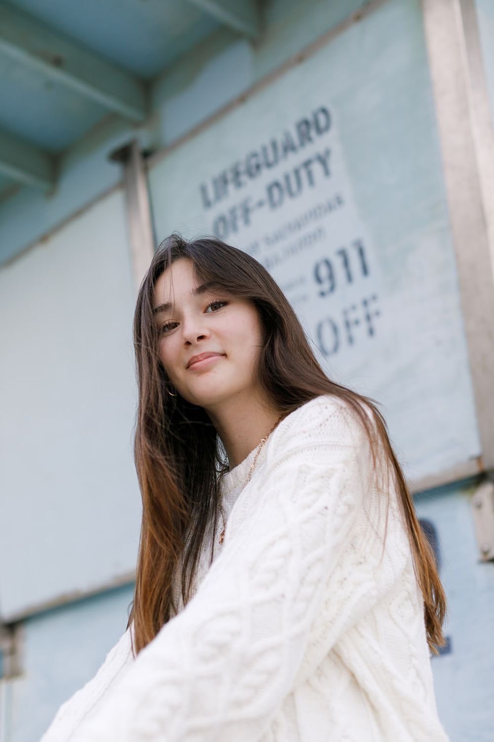 Young Asian woman with long brown hair smiles in a white knit sweater near a lifeguard station.