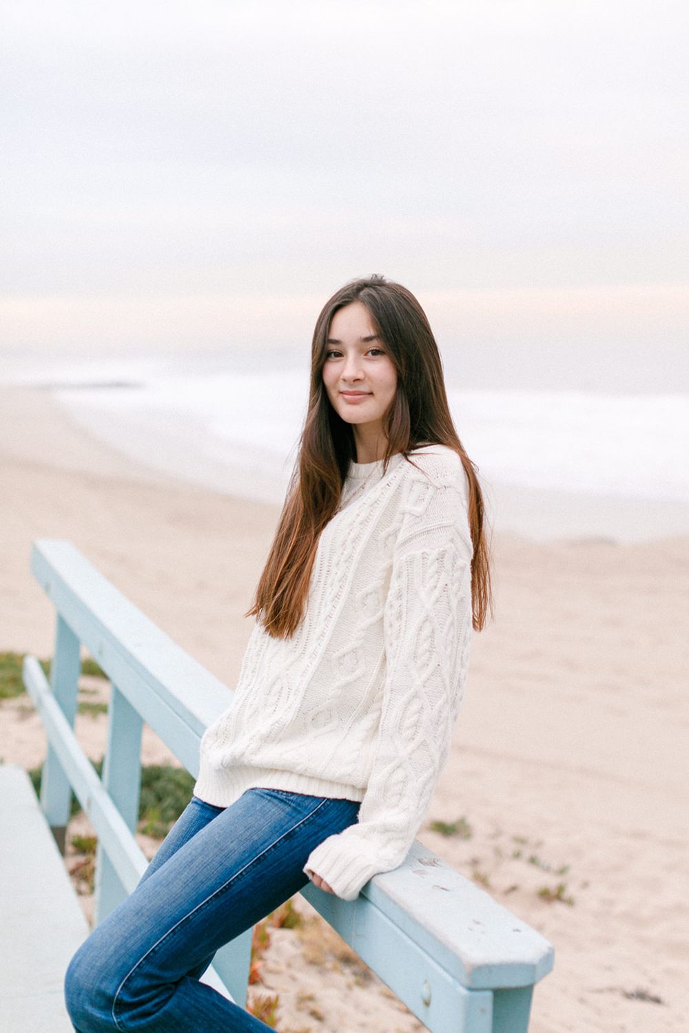 Young woman in white cable-knit sweater and jeans leans on a beach railing with ocean backdrop.