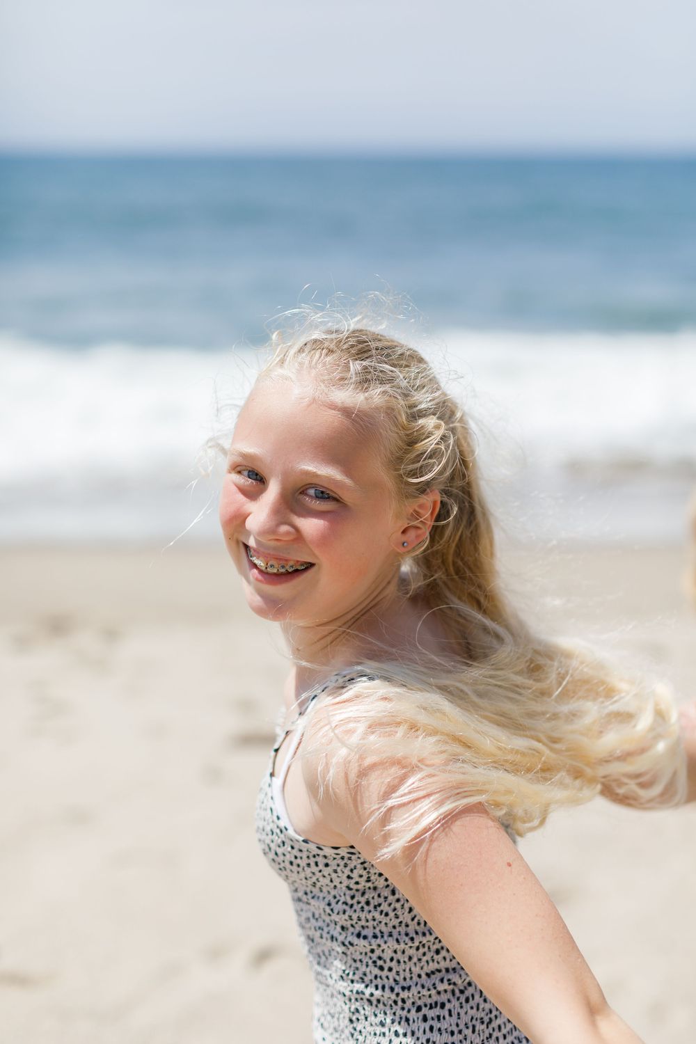 Happy blonde girl with braces laughs on a sunny beach with ocean waves in the background.