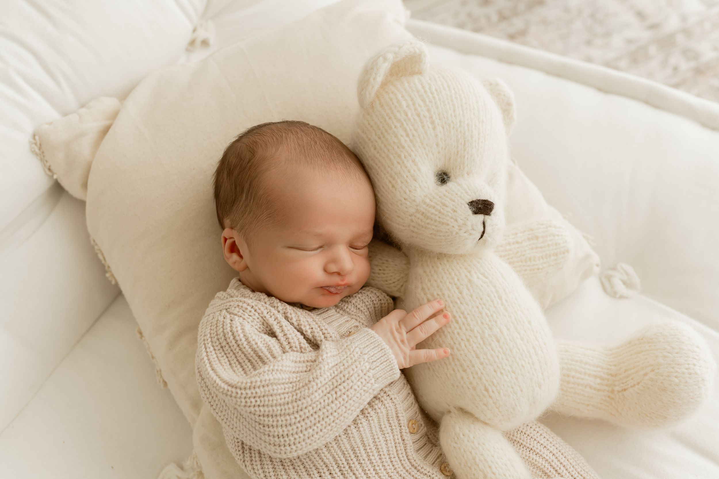 Newborn baby sleeping peacefully in white bassinet cuddling a cream knit teddy bear, wearing a cozy beige sweater.