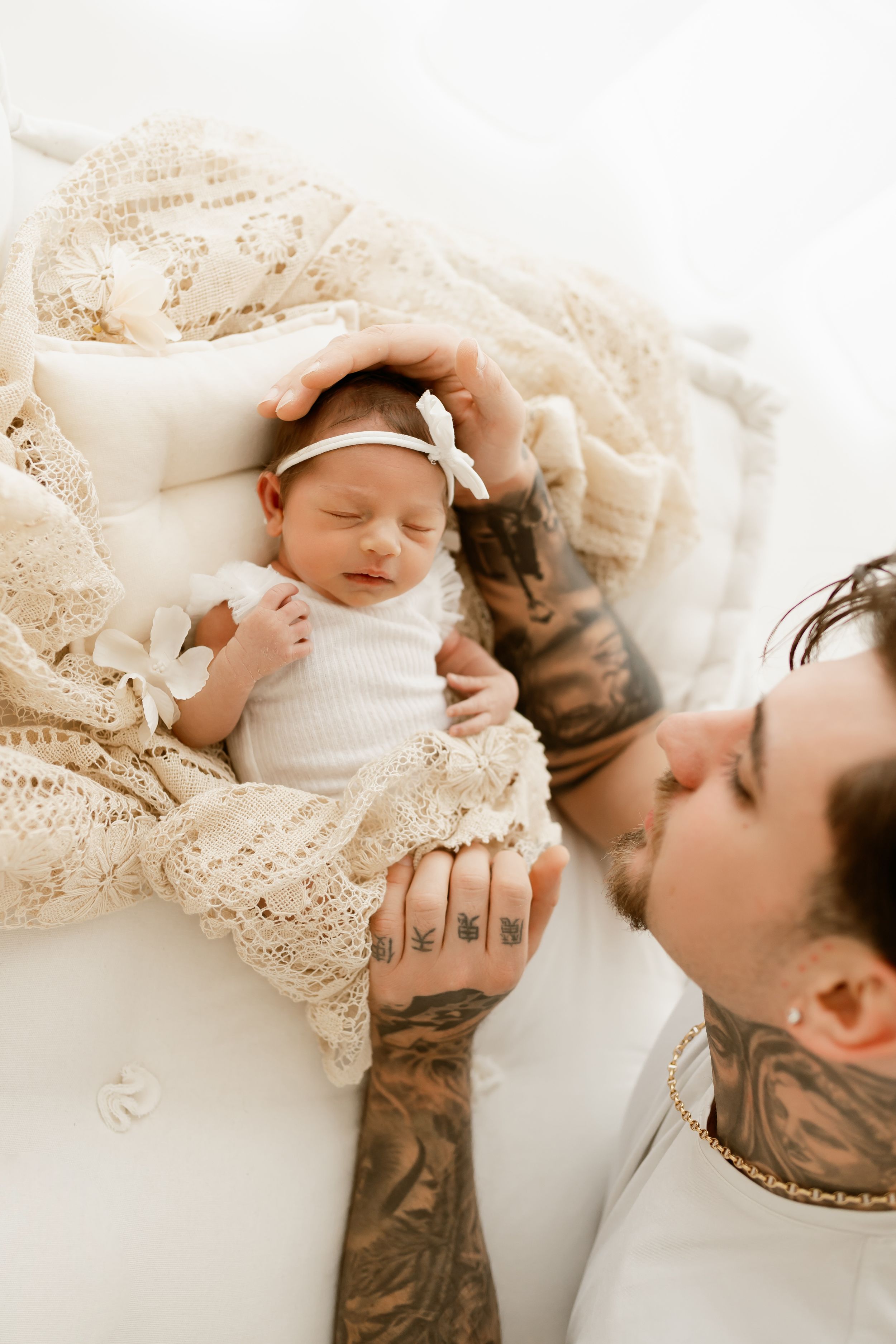 Tattooed father tenderly holds sleeping newborn baby dressed in white with bow headband on lace blanket.