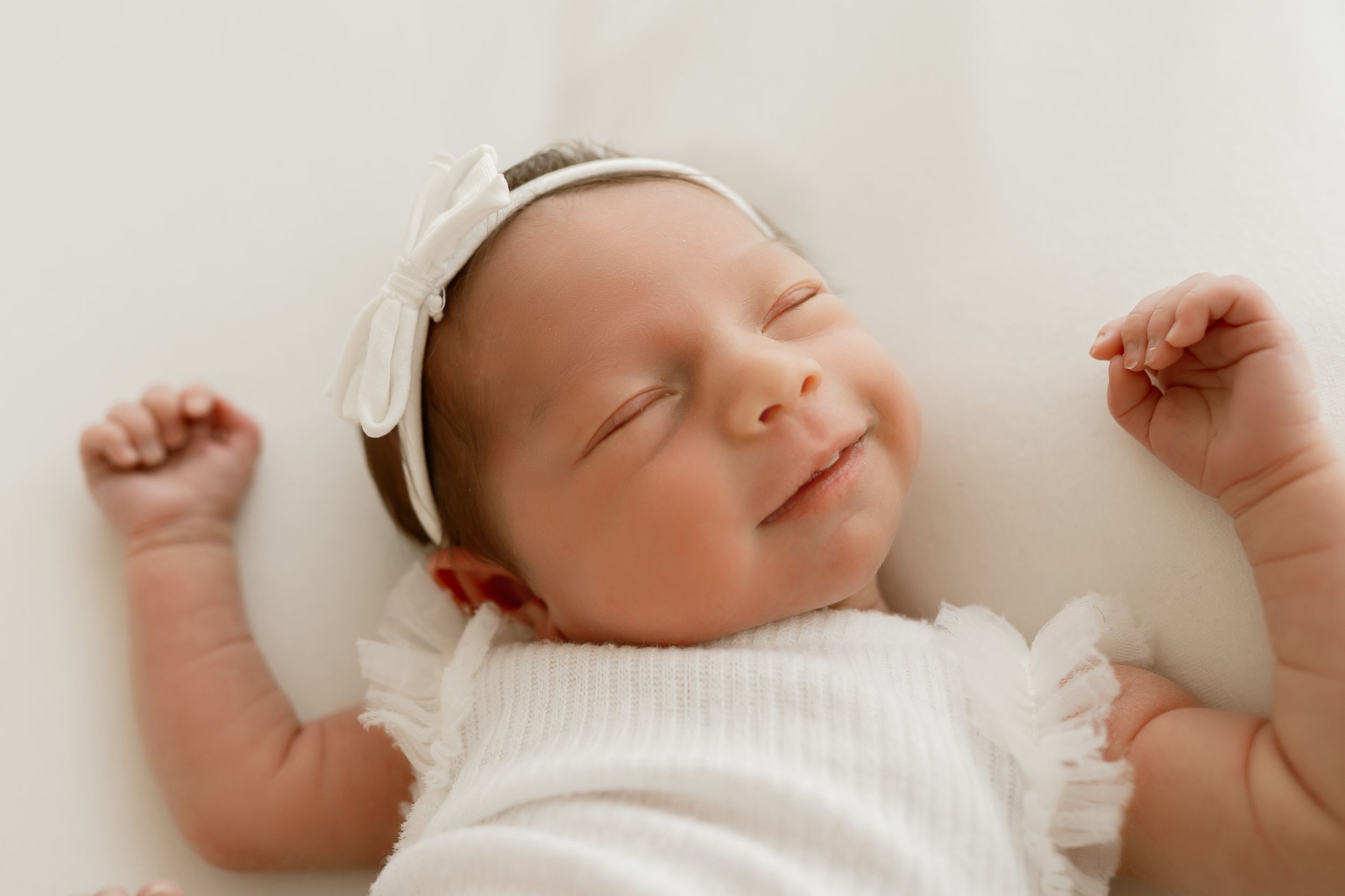 Smiling newborn baby girl wearing white headband and outfit, peacefully sleeping with arms raised.