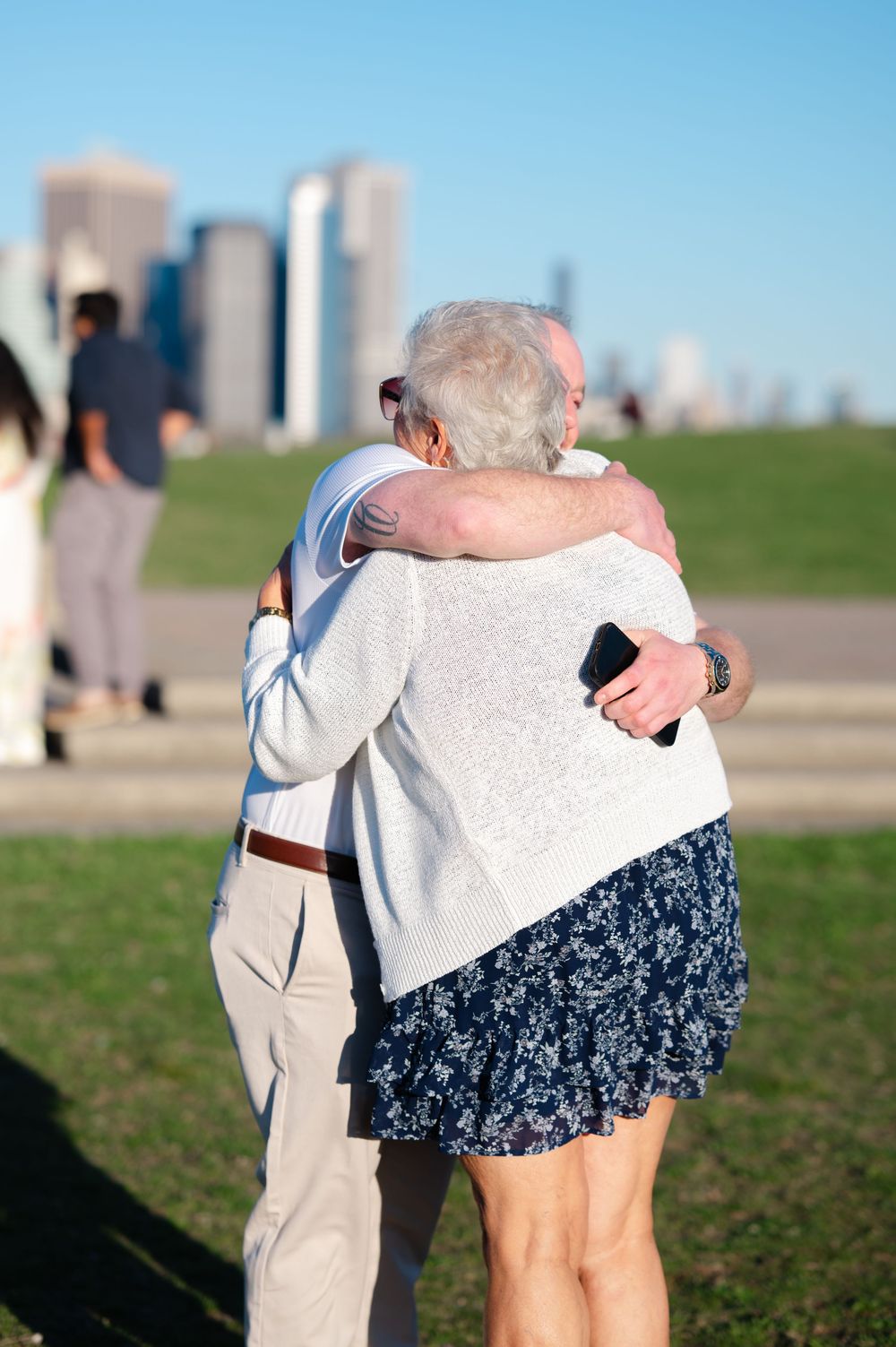 Elderly couple sharing a warm embrace outdoors with a city skyline in the background on a sunny day.
