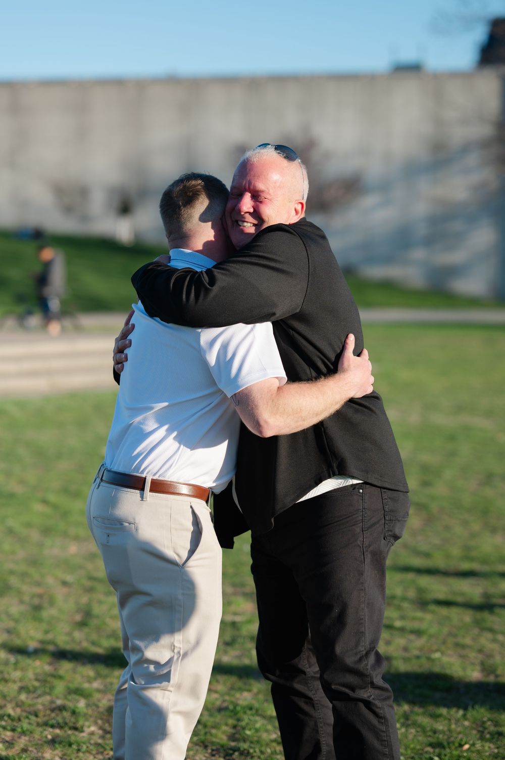 Two men share a warm embrace outdoors on a sunny day near a concrete wall.