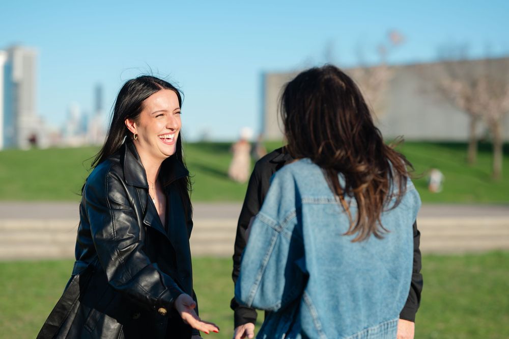 Two women smiling and shaking hands outdoors in a park with a city skyline in the background.