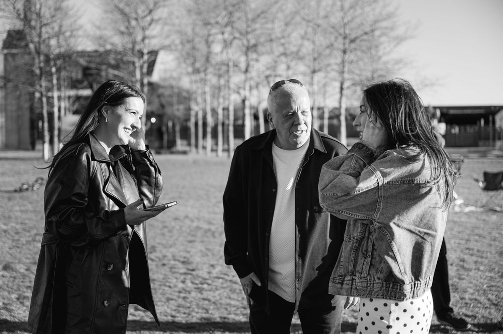 Three people conversing outdoors in a park, black and white photo, bare trees in background.