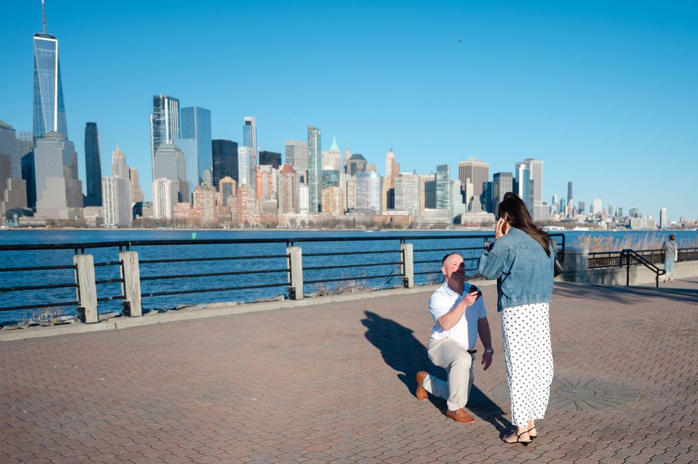 Man proposing to woman on waterfront promenade with Manhattan skyline and One World Trade Center in background.