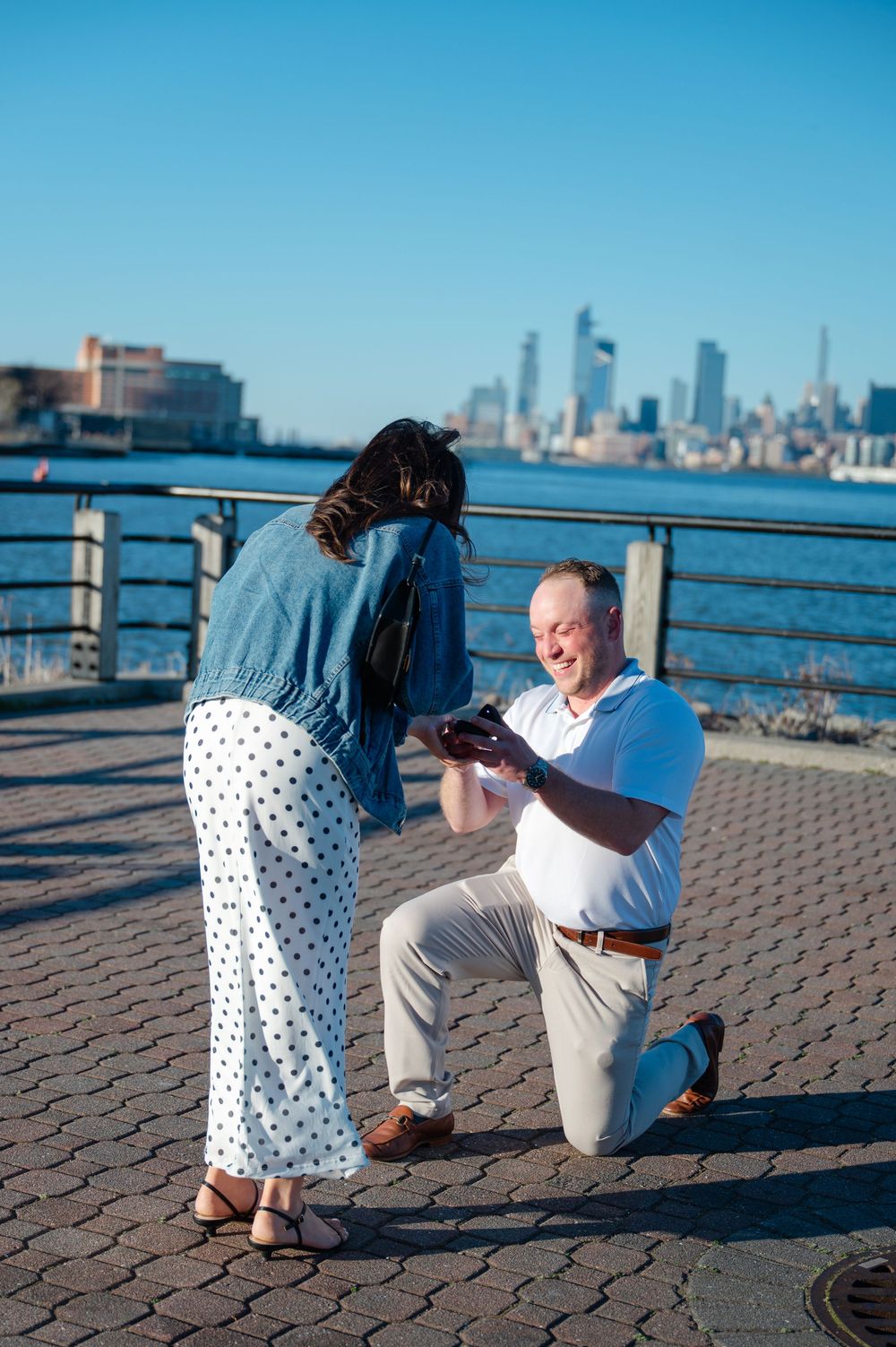 Man proposing on one knee to woman on waterfront boardwalk with NYC skyline in background.