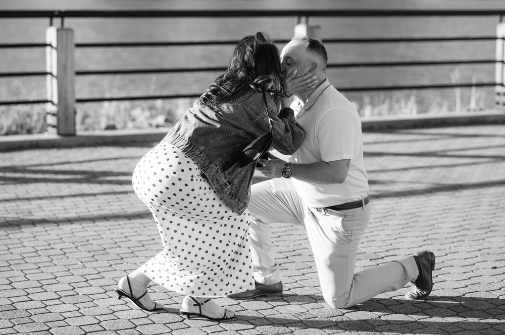 Black and white photo of a man on one knee proposing to a woman on a sunny brick plaza.