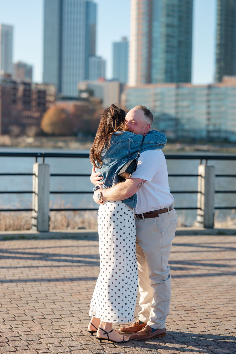 Couple embracing and kissing on a waterfront promenade with city skyline in the background.