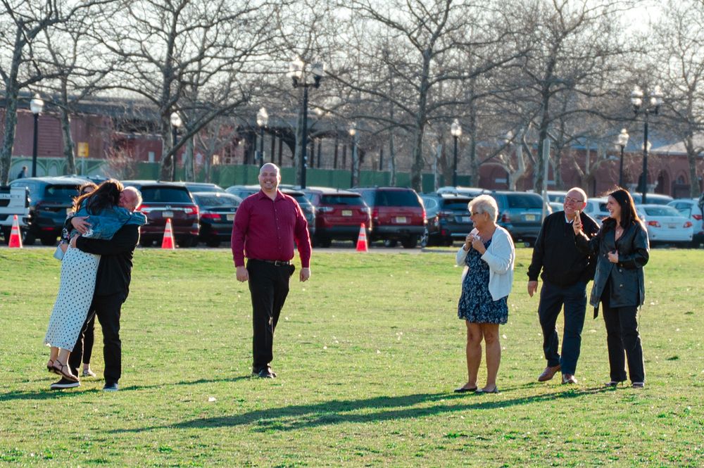 Group of adults socializing on a green lawn near a parking lot with bare trees on a sunny day.