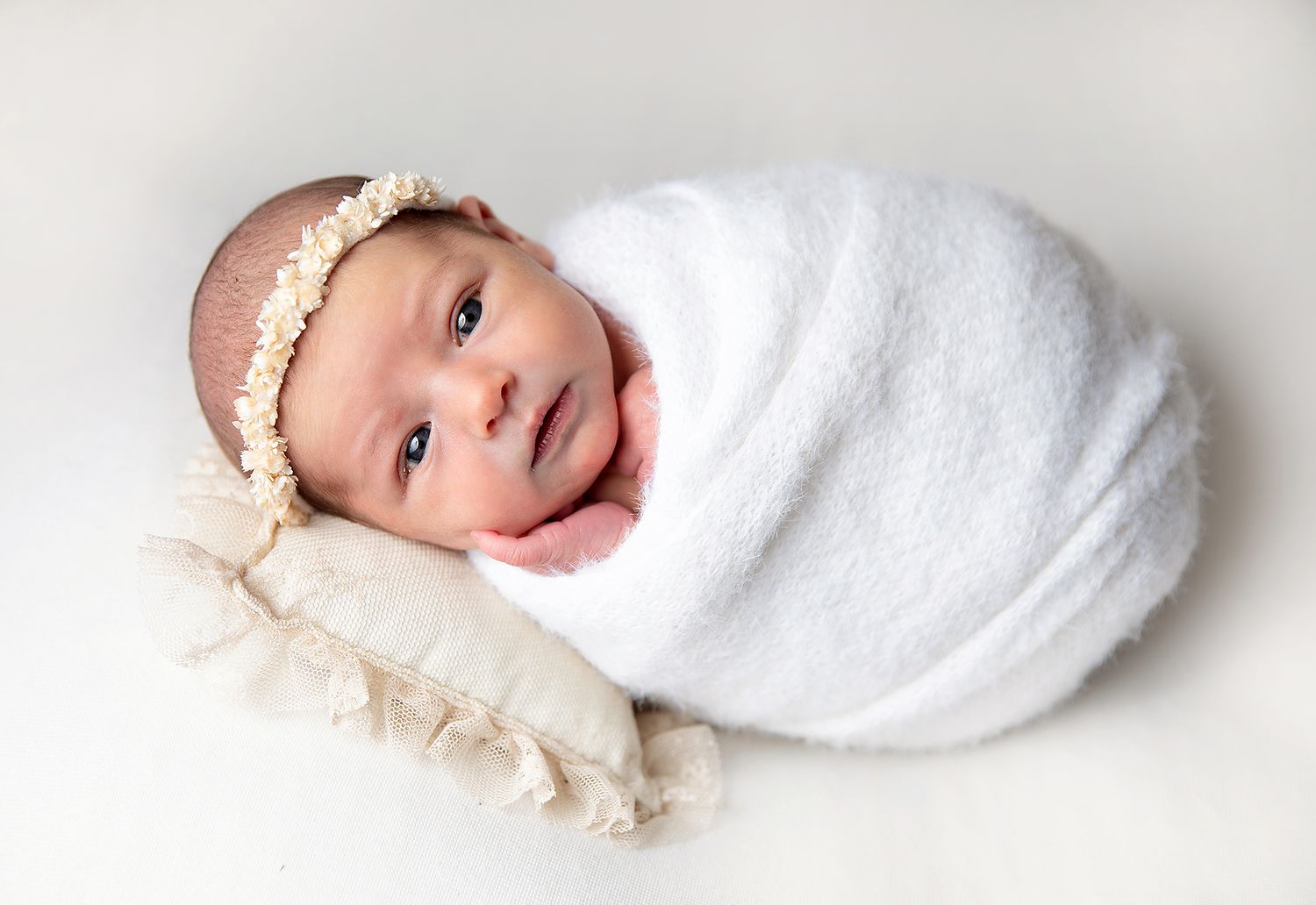 Newborn baby swaddled in white wrap with floral headband, resting on cream pillow. During a newborn session in South East London