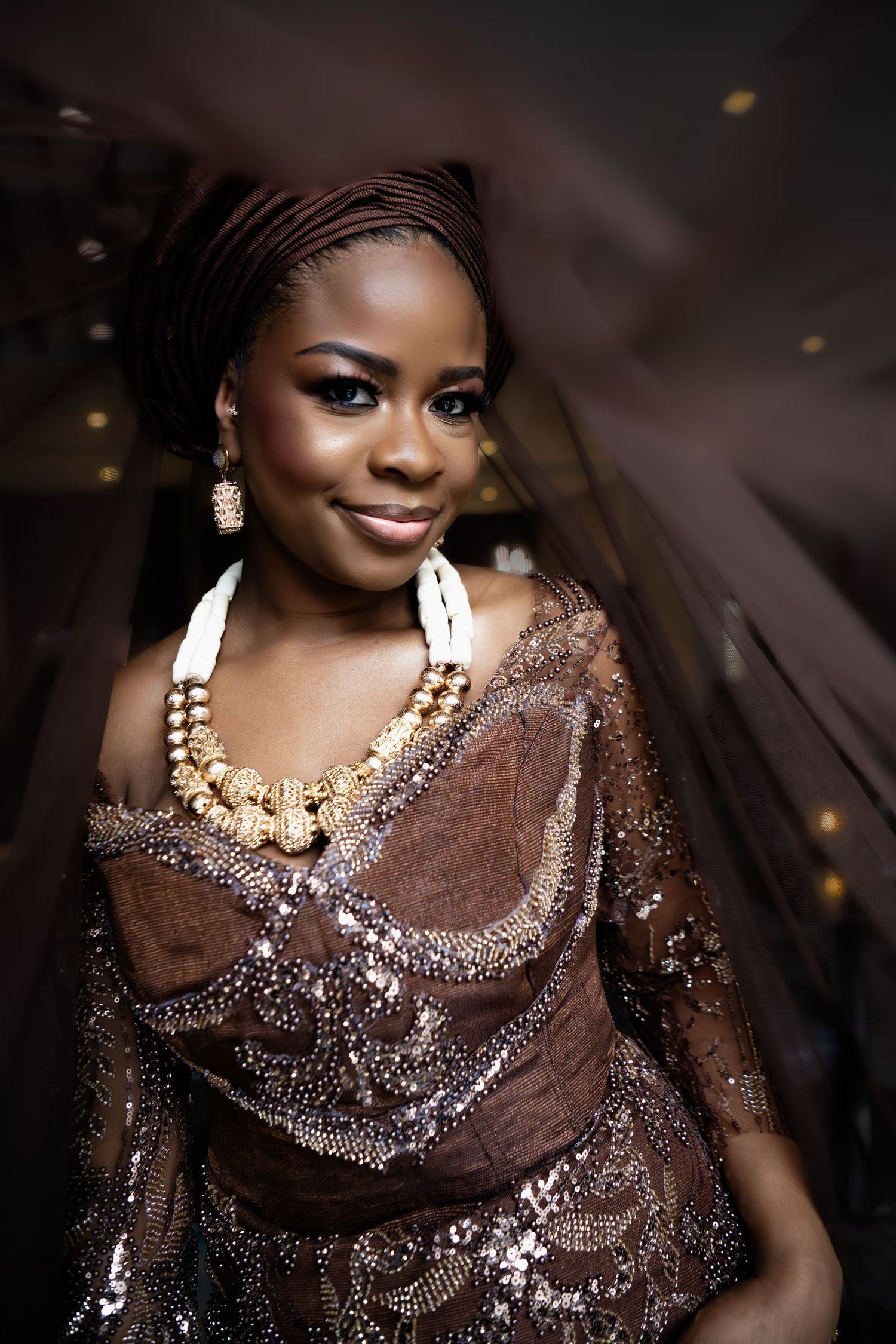 Elegant bride in ornate brown beaded gown with matching headwrap and gold jewelry at a Nigerian wedding.