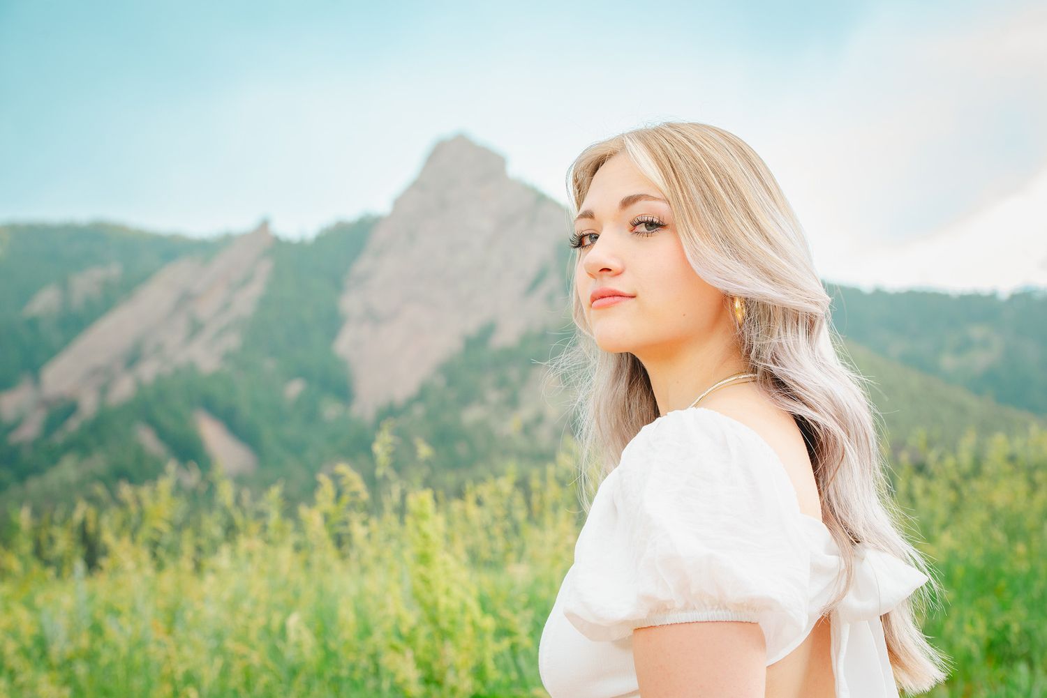 Young woman with blonde hair standing in a lush green field with mountains in the background.