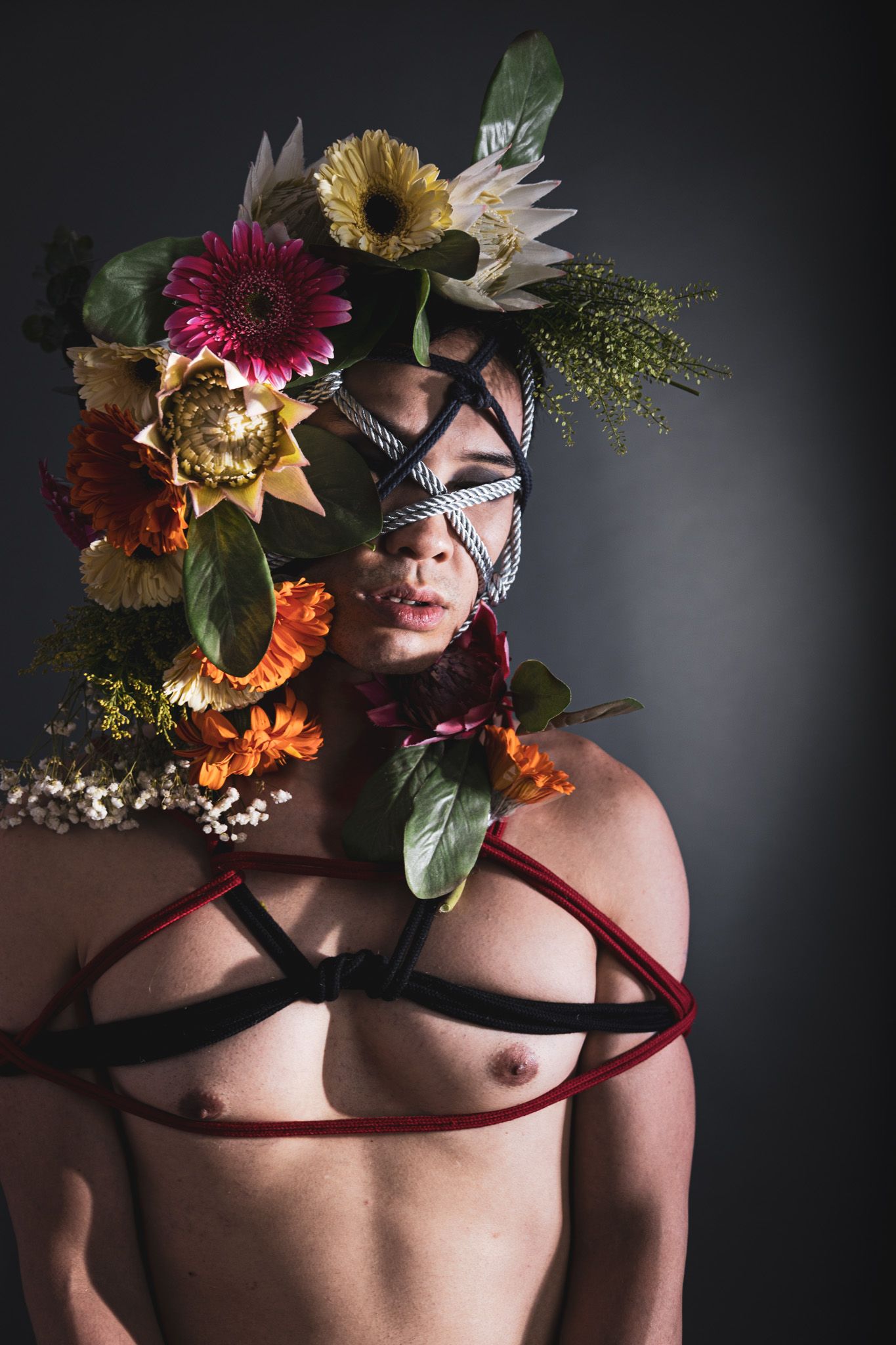 Shirtless person with colorful floral headpiece and intricate rope face and chest bondage harness.
