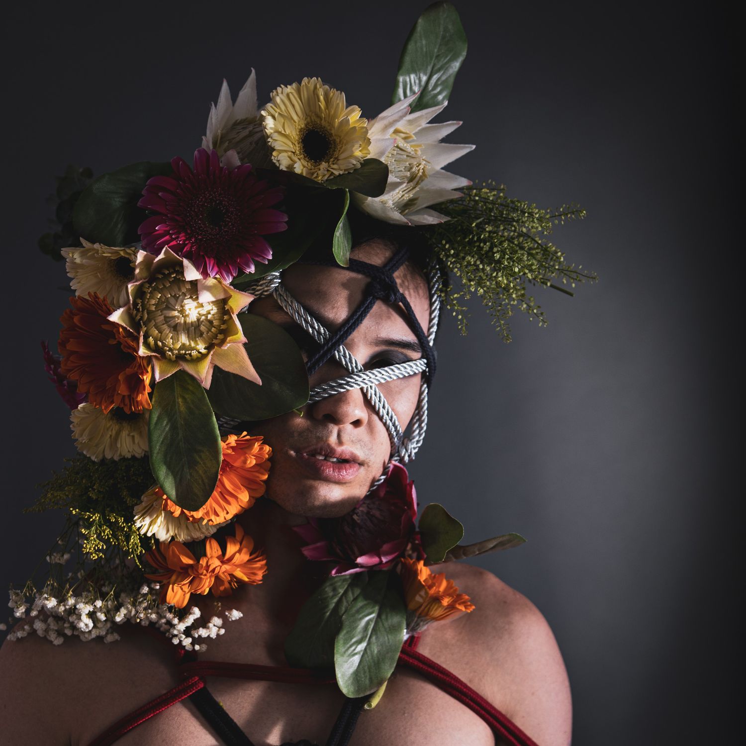 Close-up portrait of person with rope face bondage adorned with colorful flowers including gerberas.