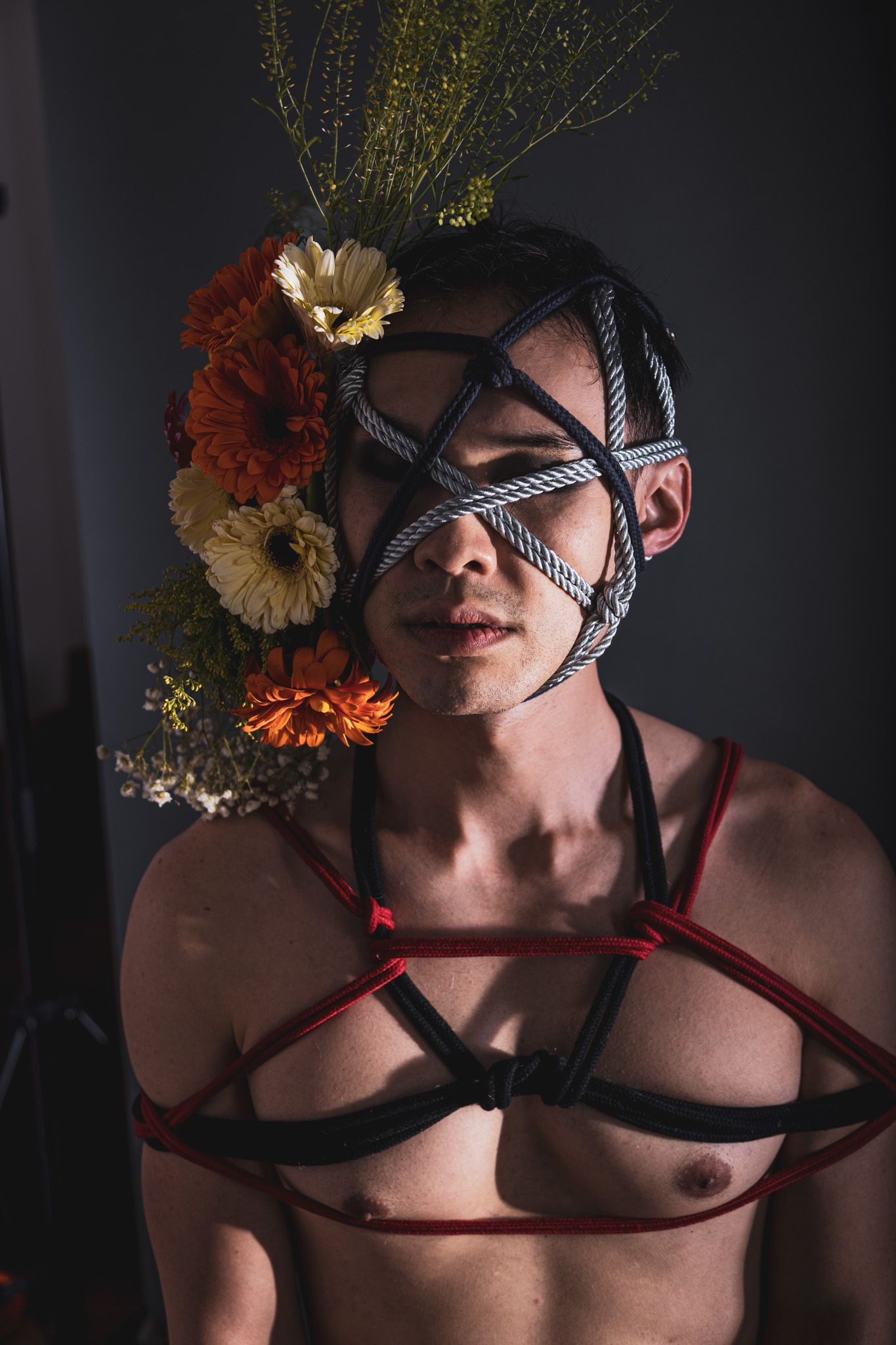 Shirtless person with rope face bondage and small floral arrangement, wearing red and black harness.
