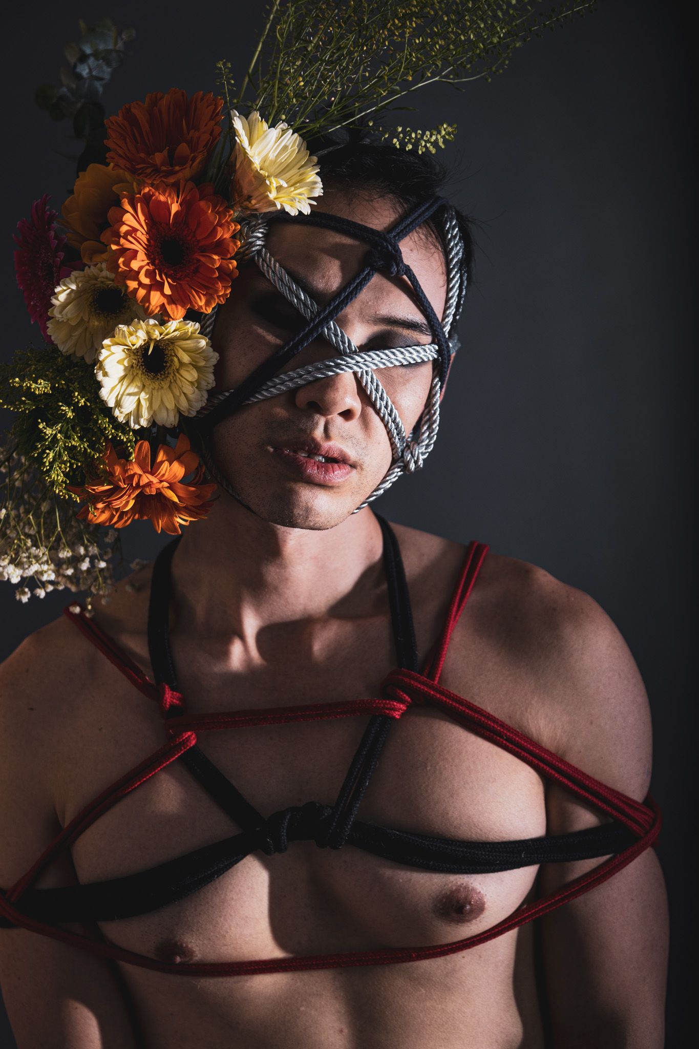 Shirtless person with rope face bondage decorated with orange and yellow flowers and red harness.
