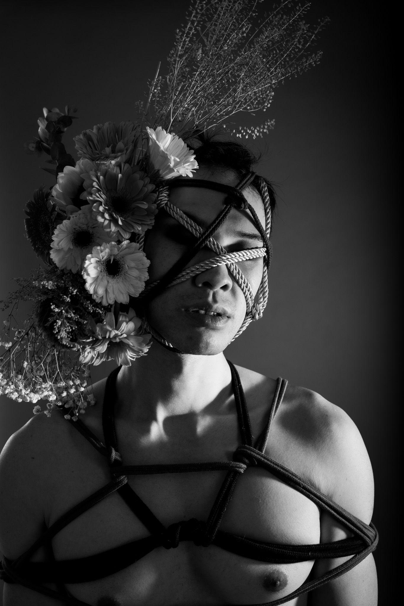 Black and white portrait of person with rope face bondage adorned with flowers and black harness.