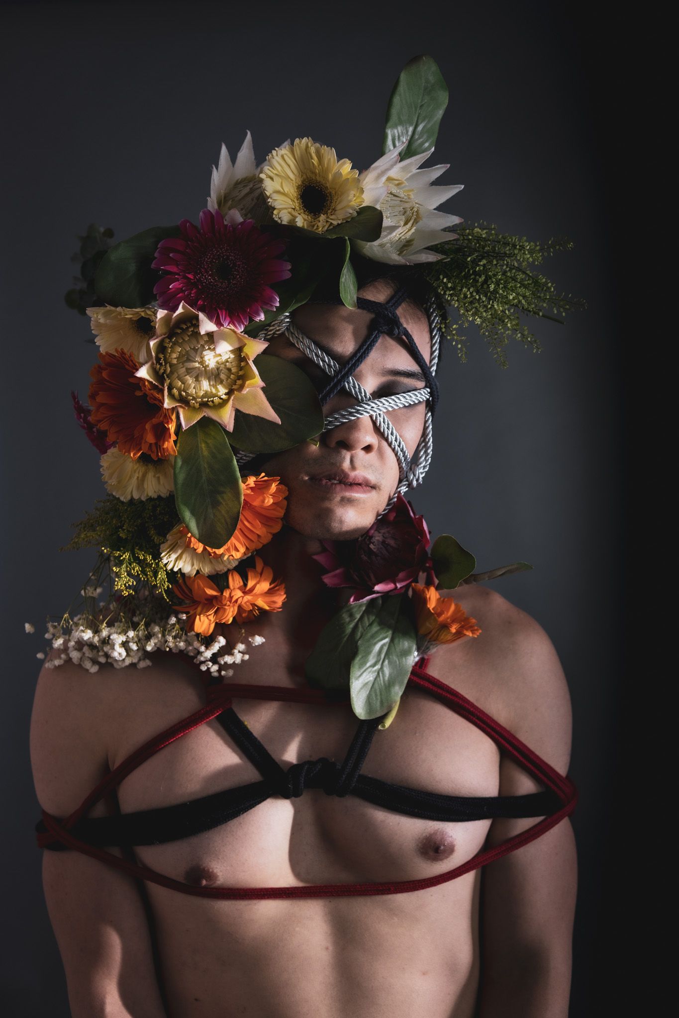 Person with elaborate floral crown and rope face bondage, shirtless with red and black rope harness.