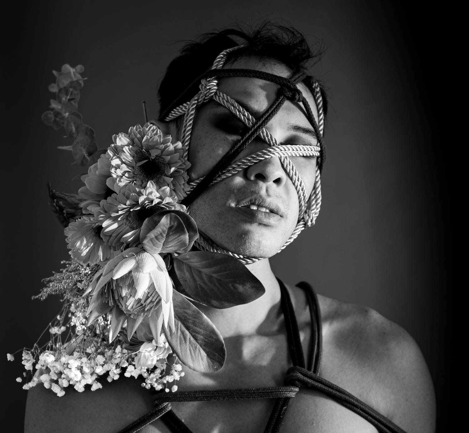 Black and white portrait of person with rope blindfold and floral arrangement with feathers and flowers.