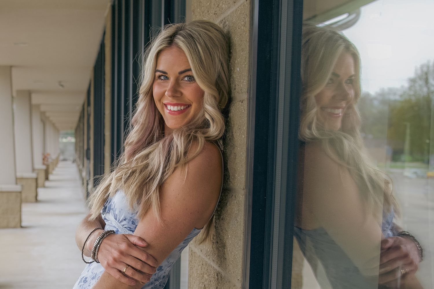Smiling blonde woman leaning against a stone pillar, reflected in glass windows along a covered walkway.