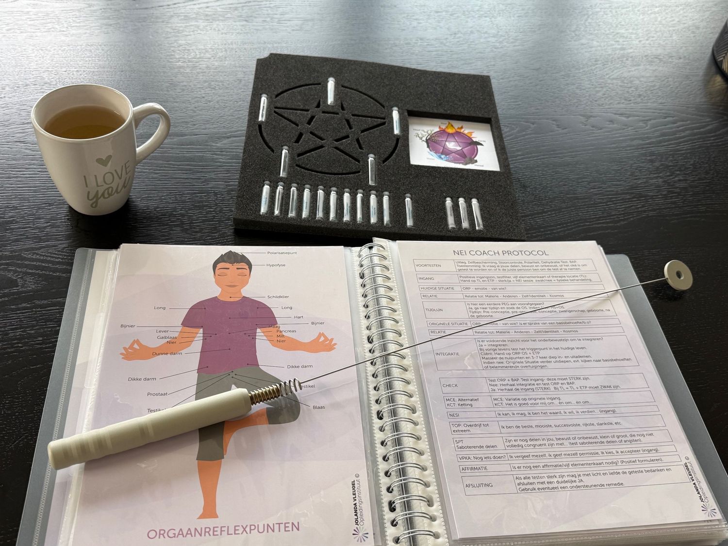 Open reflexology binder with organ reflex points diagram, pendulum, pentagram board, and tea cup on dark table.