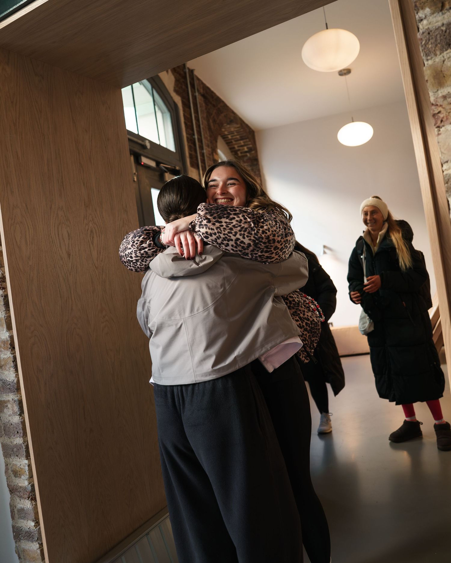 Two people share a warm hug in a modern hallway while another person stands in the background.