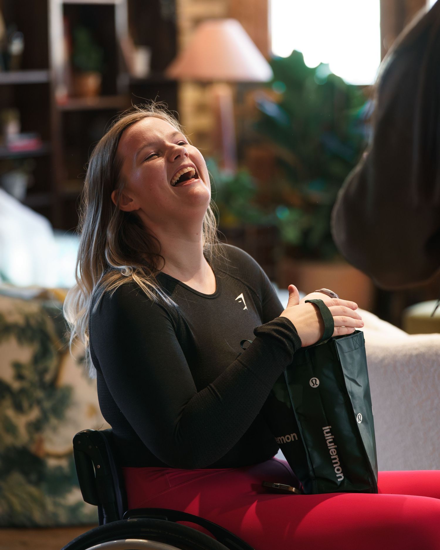 A young woman in a wheelchair laughs joyfully while holding a lululemon bag in a warmly lit living room.