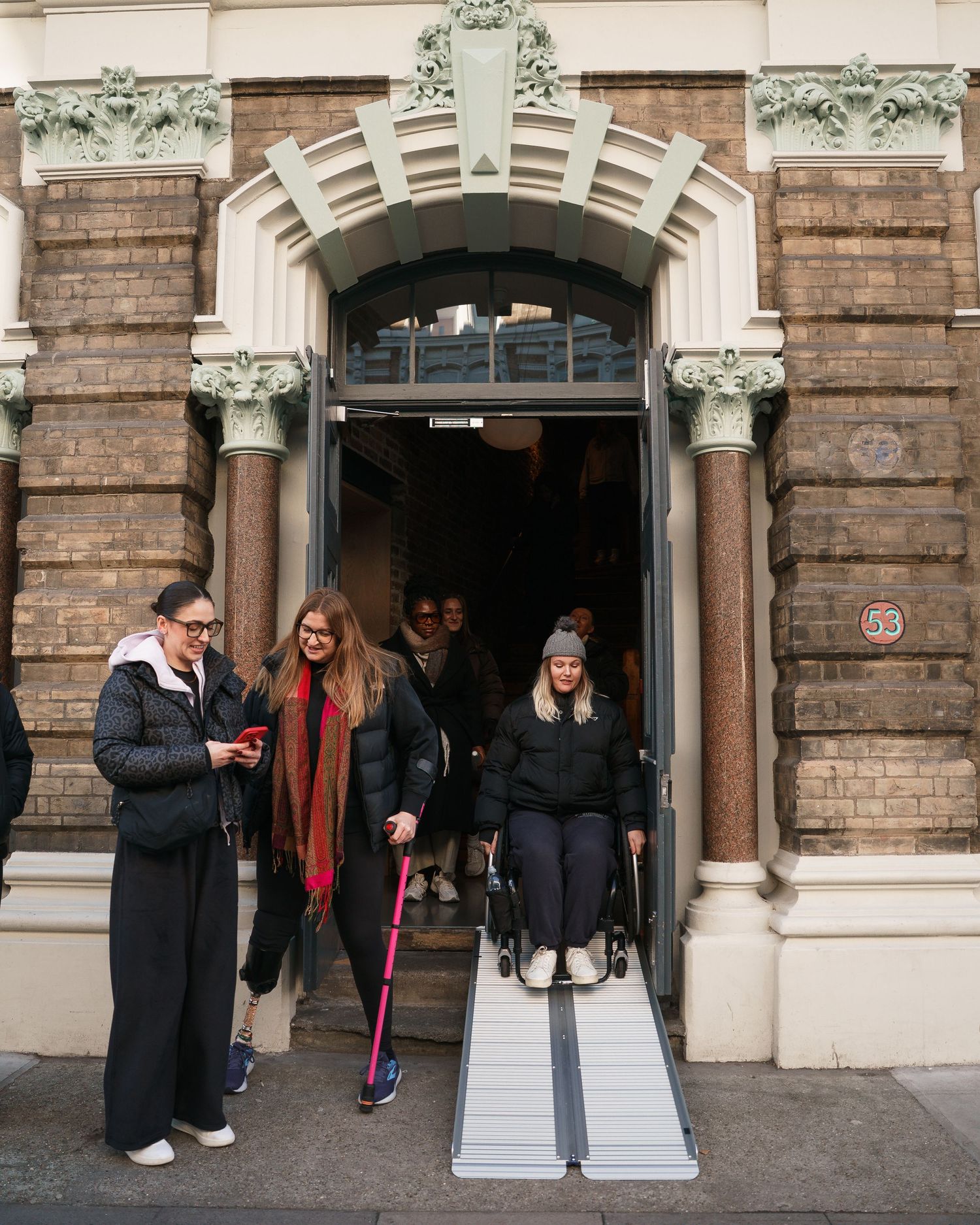 Woman in wheelchair exits historic brick building via ramp, assisted by friends on a city street.