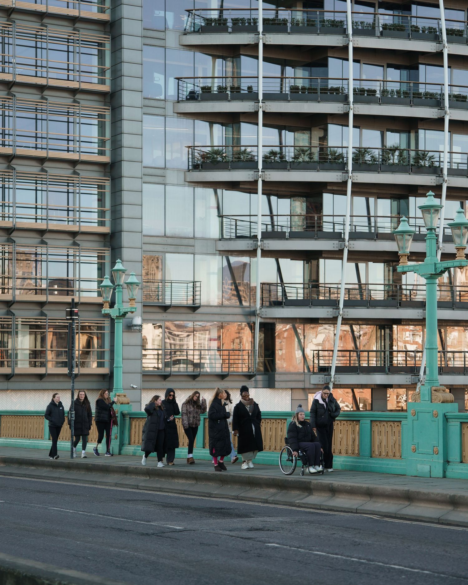People walking and a wheelchair user on a teal bridge with a modern glass apartment building in London backdrop.