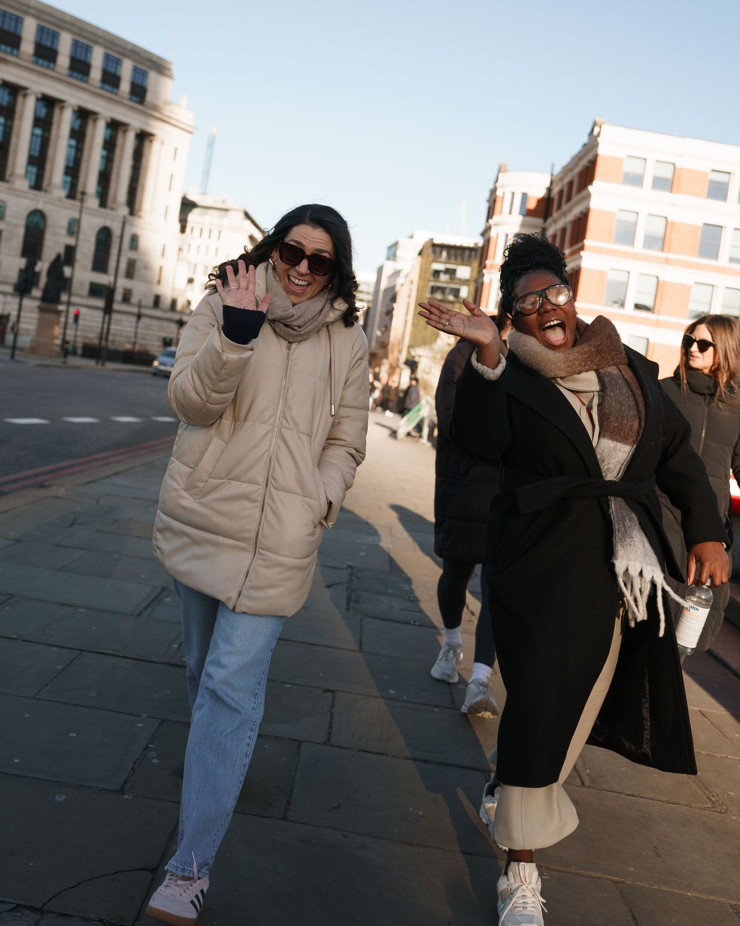 Two women posing and laughing on a city street wearing winter coats on a sunny day.
