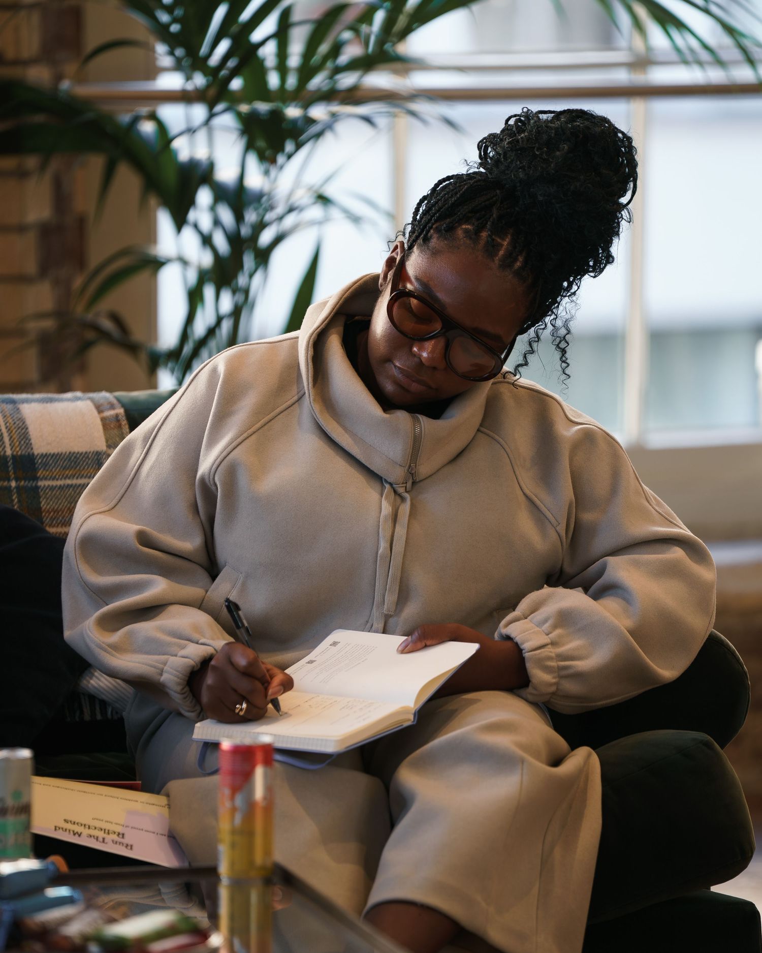 A young Black woman with braids writes in a notebook, wearing an oversized beige hoodie, seated in a cozy indoor space.