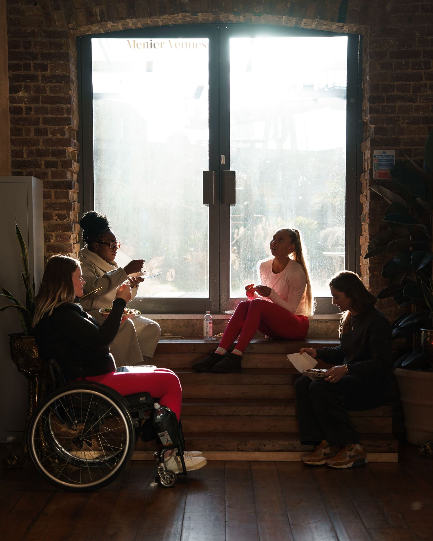 Diverse group of women socializing near a sunlit window, one in a wheelchair, in a modern brick-walled space.