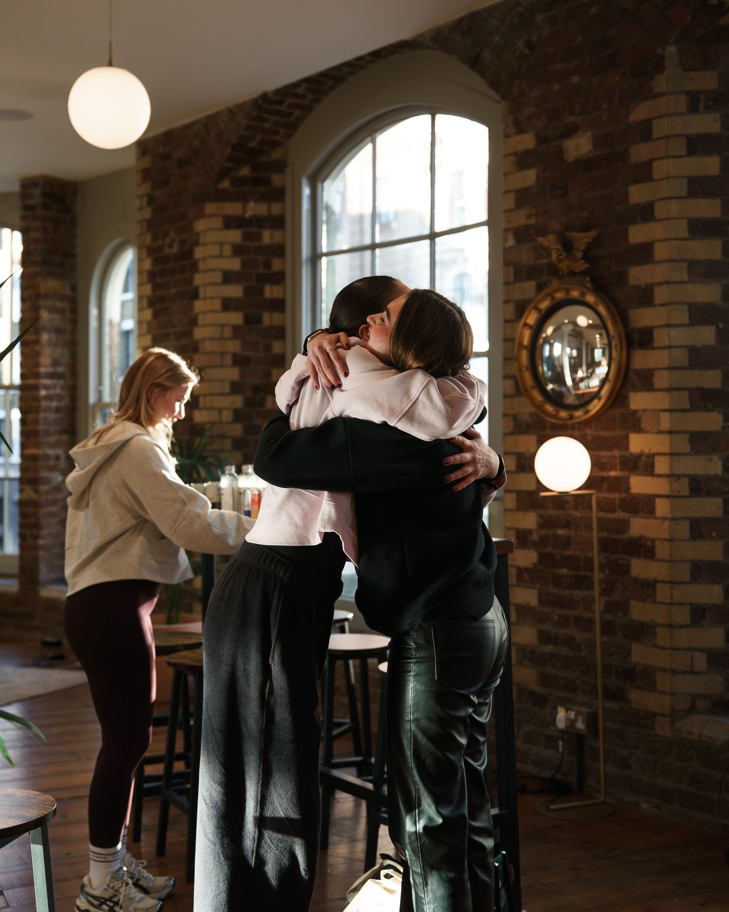 Two people share a warm hug in a cozy brick-walled cafe while a woman stands at the bar nearby.