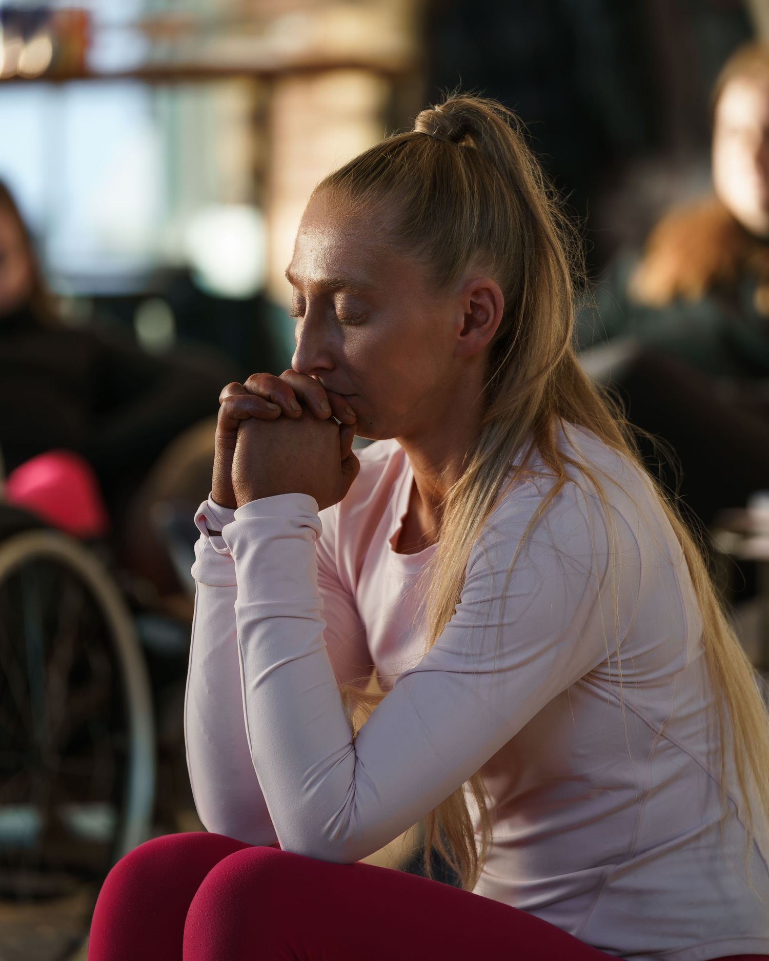 Blonde woman in pink athletic wear sitting with hands clasped in a contemplative pose.