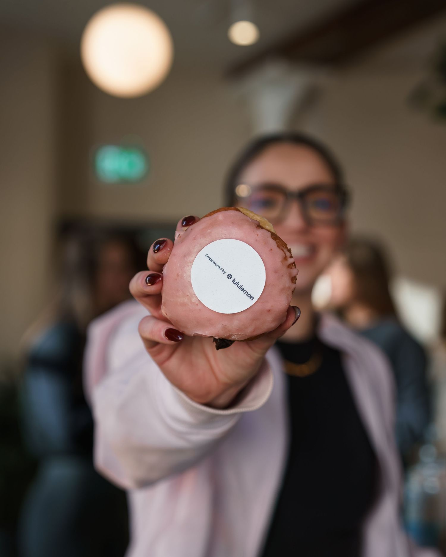 Woman holds up a branded cupcake with a white logo sticker toward the camera in a cafe.