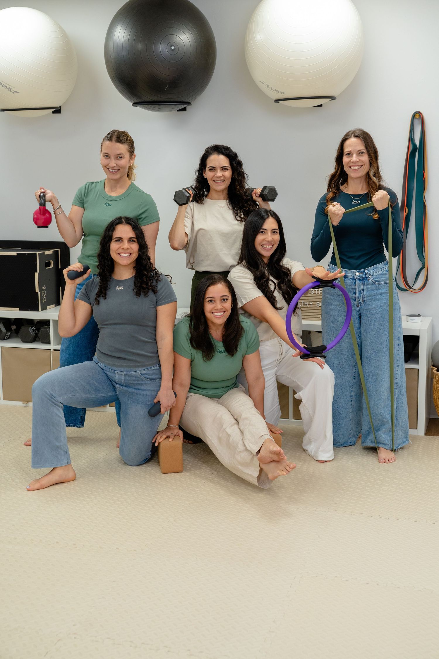 Six women pose in a fitness studio holding dumbbells, a pilates ring, and resistance bands with exercise balls in background.
