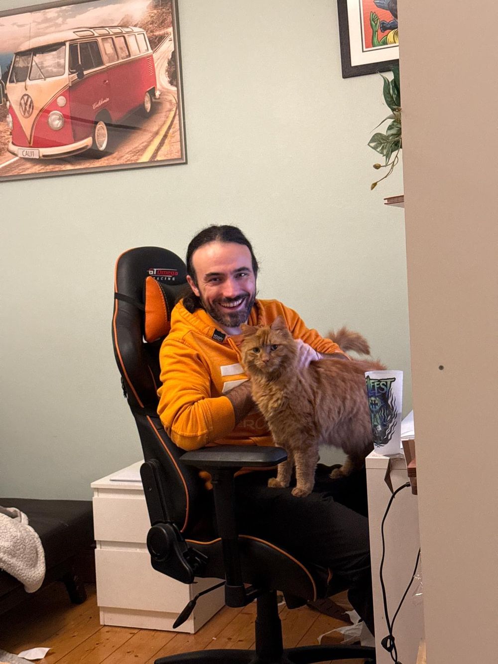Smiling man in orange hoodie sits in gaming chair holding a fluffy ginger cat in a cozy room.