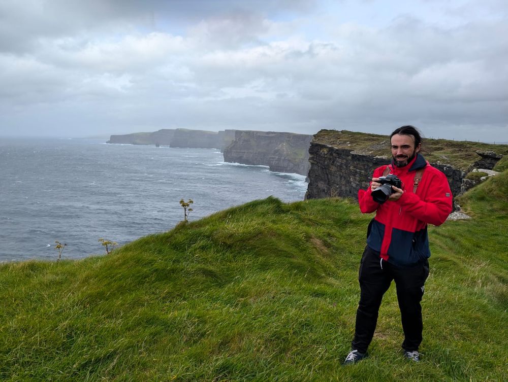 Man holding camera at Cliffs of Moher Ireland, wearing red jacket, dramatic coastal cliffs and ocean in background.