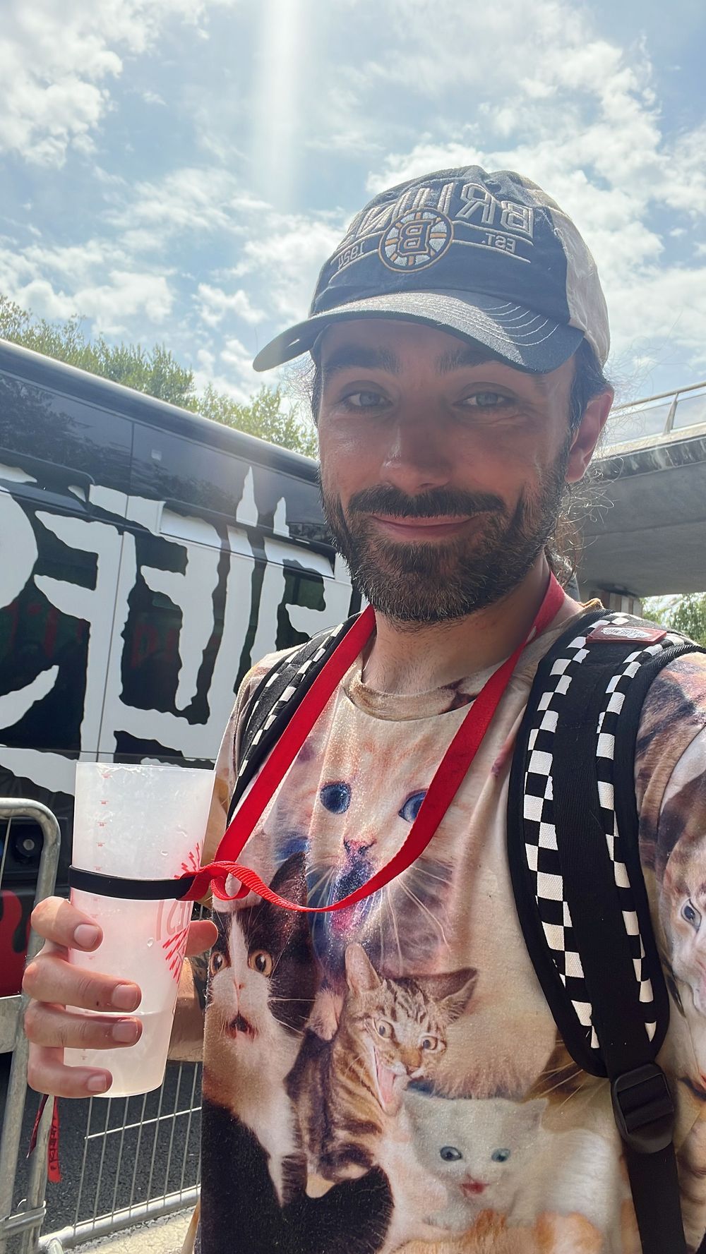 Bearded man wearing a cat-print shirt and baseball cap holds a drink at an outdoor festival.
